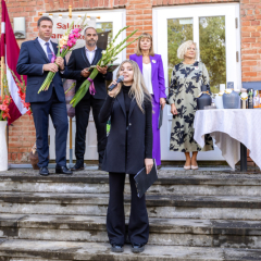 Meitene runā dzejoli, fonā skolas padomes priekšsēdētājs, pašvaldības pārstāvis, skolas direktore un mācību pārzine.